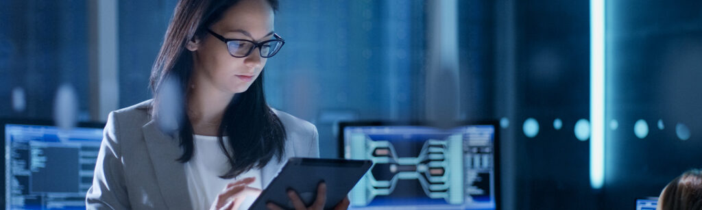 Young Female Government Employee Wearing Glasses Uses Tablet in System Control Center. In the Background Her Coworkers are at Their Workspaces with many Displays Showing Valuable Data.